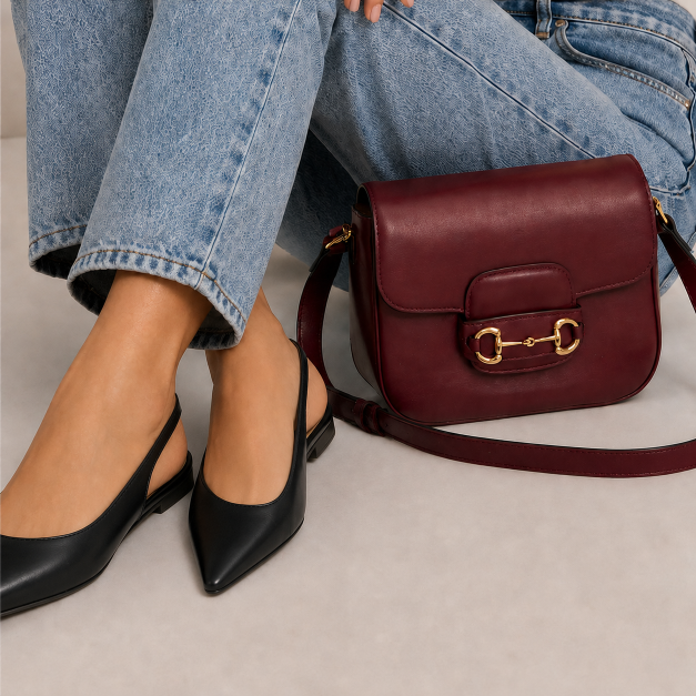 A burgundy leather shoulder bag with a gold horsebit clasp, styled next to a person wearing light-wash blue jeans and black pointed slingback flats on a neutral beige floor.
