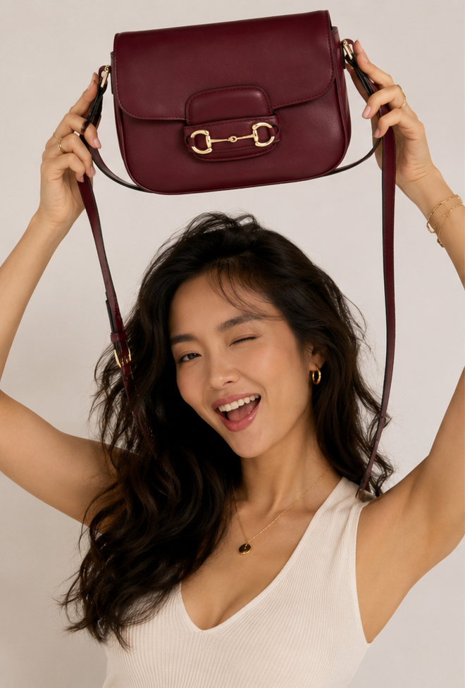 A woman with long dark hair holds a burgundy leather shoulder bag above her head. She is wearing a white ribbed tank top and light blue denim jeans against a plain light gray background.