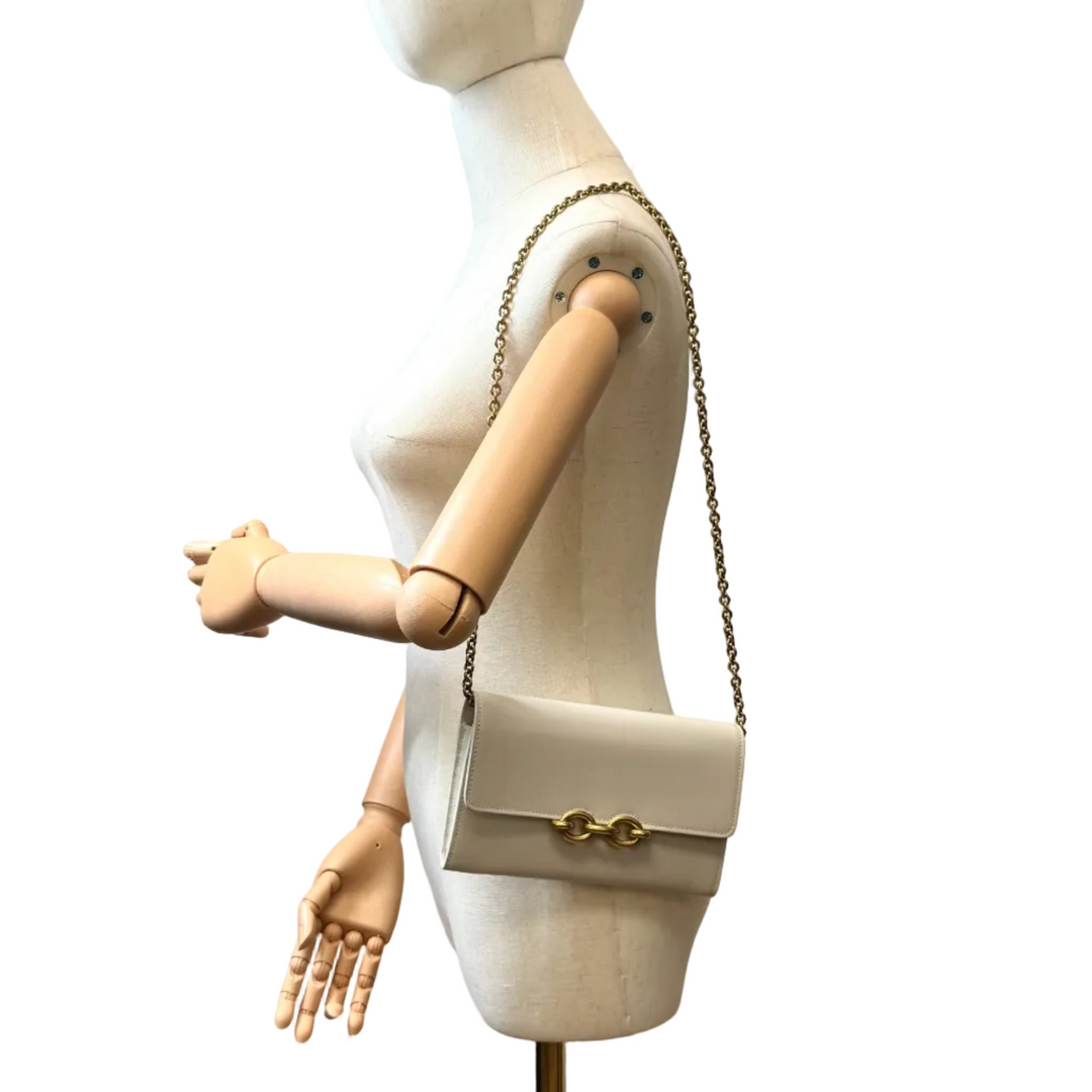 A cream-colored leather shoulder bag with a gold chain strap and double gold clasp, displayed on a cream mannequin torso. The mannequin is set against a white wall with a colorful painting and a person's arm pointing at it.