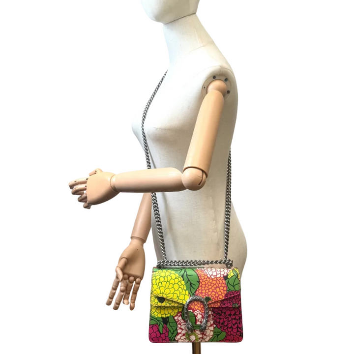 A colorful crossbody handbag with a chain strap, displayed on a wooden mannequin torso against a white background. The bag features a vibrant, multicolored pattern of stylized fruits (yellow, orange, green, and pink) with a prominent gold-tone 'G' logo clasp on the front.