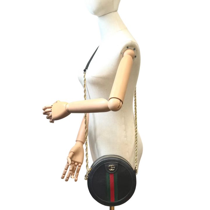 A black round shoulder bag with a gold chain strap and a red vertical stripe, displayed on a white mannequin torso in an indoor room with a white wall, colorful painting, and wooden floor.