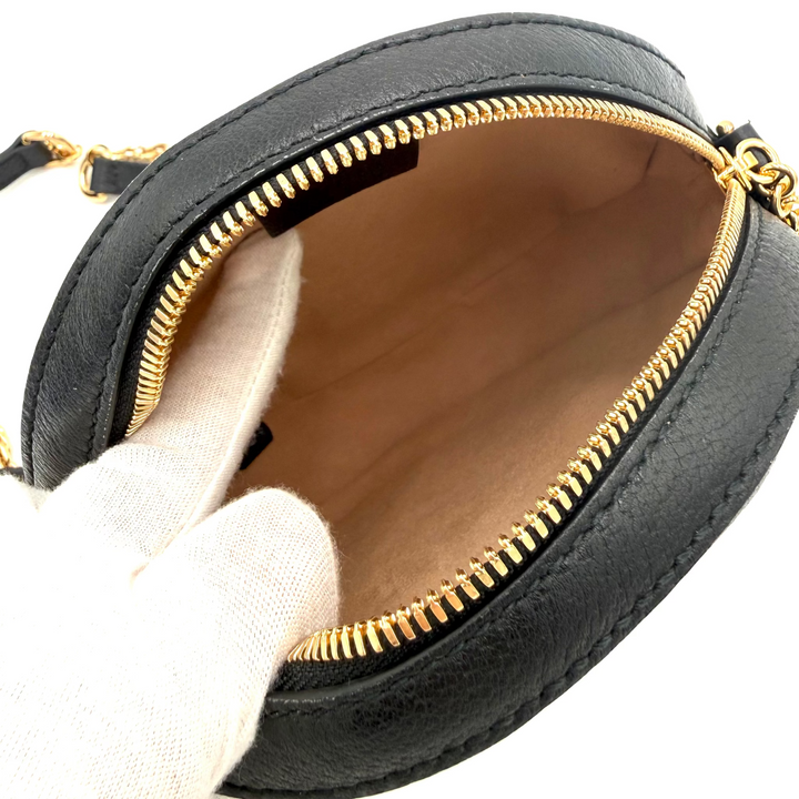 A close-up of a black leather crossbody bag with gold-tone hardware, shown open with a white gloved hand holding the flap to display the interior lining.