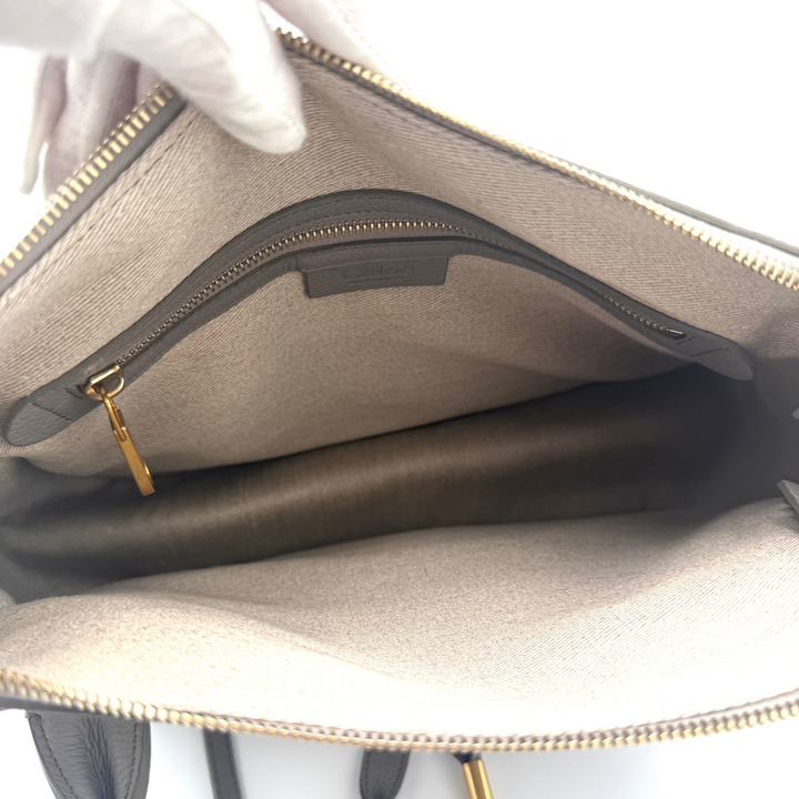 An open beige handbag shown from the inside, featuring a main zip closure, a zippered pocket, and gold-tone hardware. A white-gloved hand is holding the bag open against a plain white background.