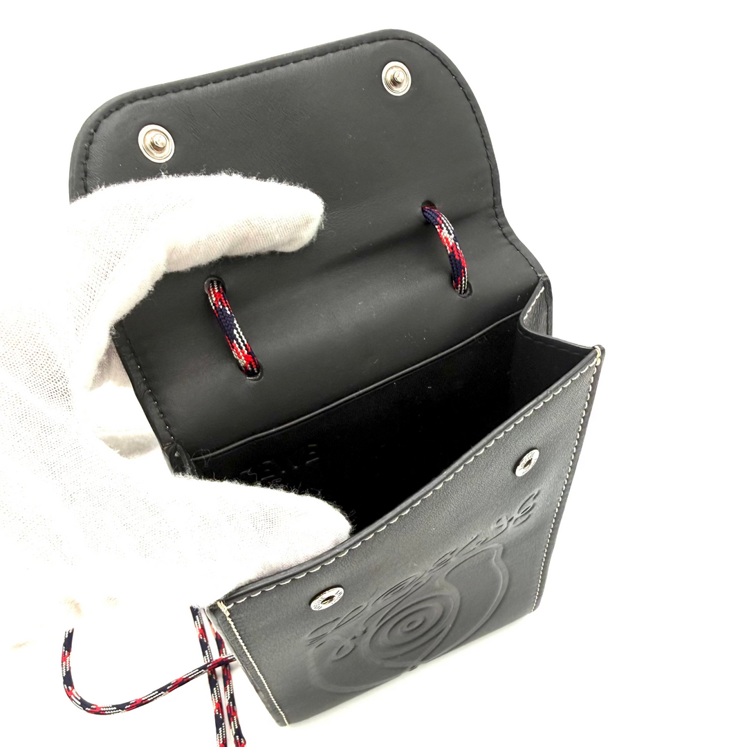 A black leather pouch with a spiral design embossed on the front, held open by a white-gloved hand. The pouch features silver-tone metal snap buttons, a red and black braided lanyard, and a black leather strap with a knot, shown against a plain white background.