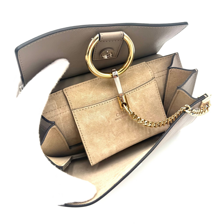 A structured shoulder bag in taupe leather with a large gold-tone O-ring handle and a gold chain strap, shown open against a white background.