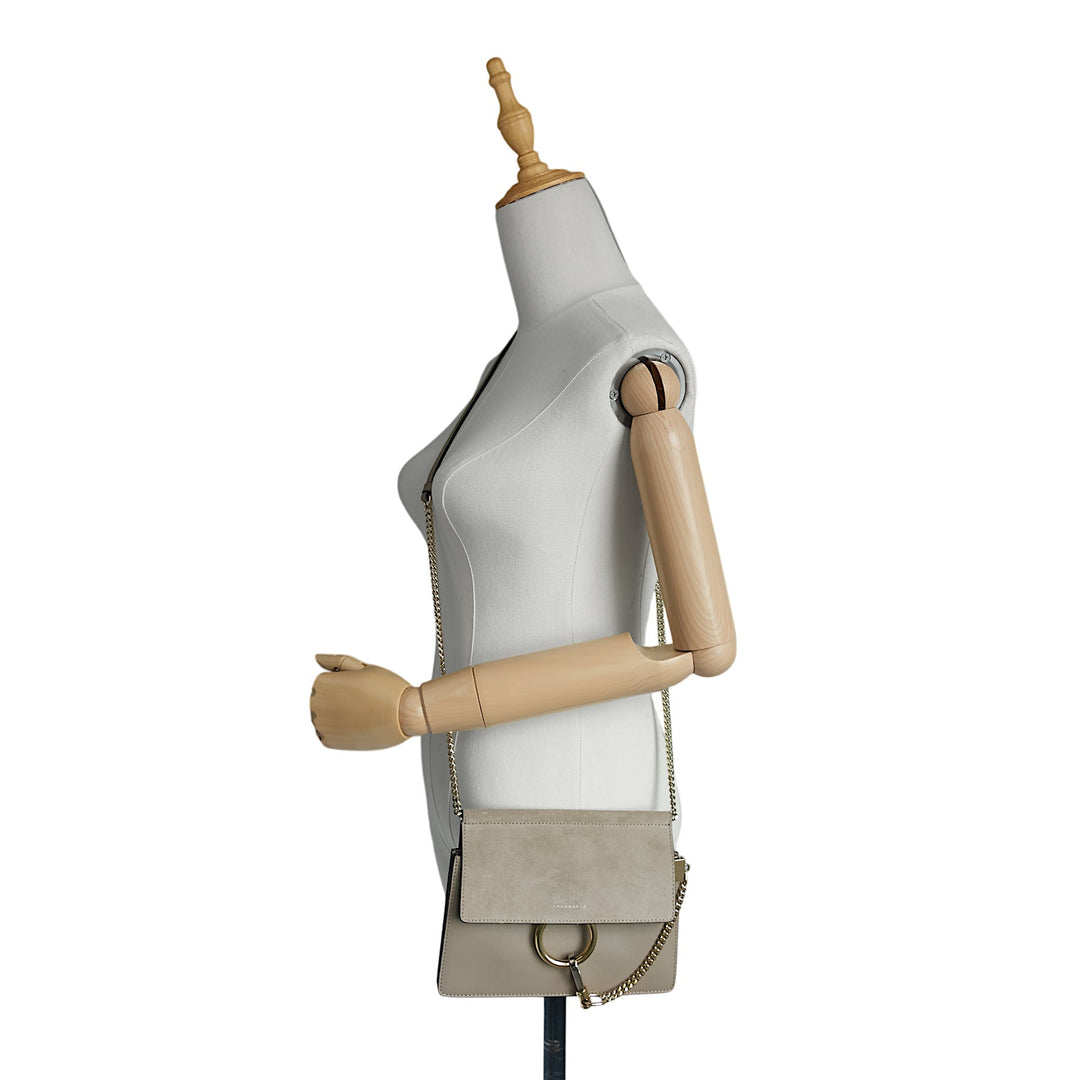 A light beige leather shoulder bag with a flap closure and gold-tone chain strap, displayed on a white mannequin torso with wooden arms against a white background.